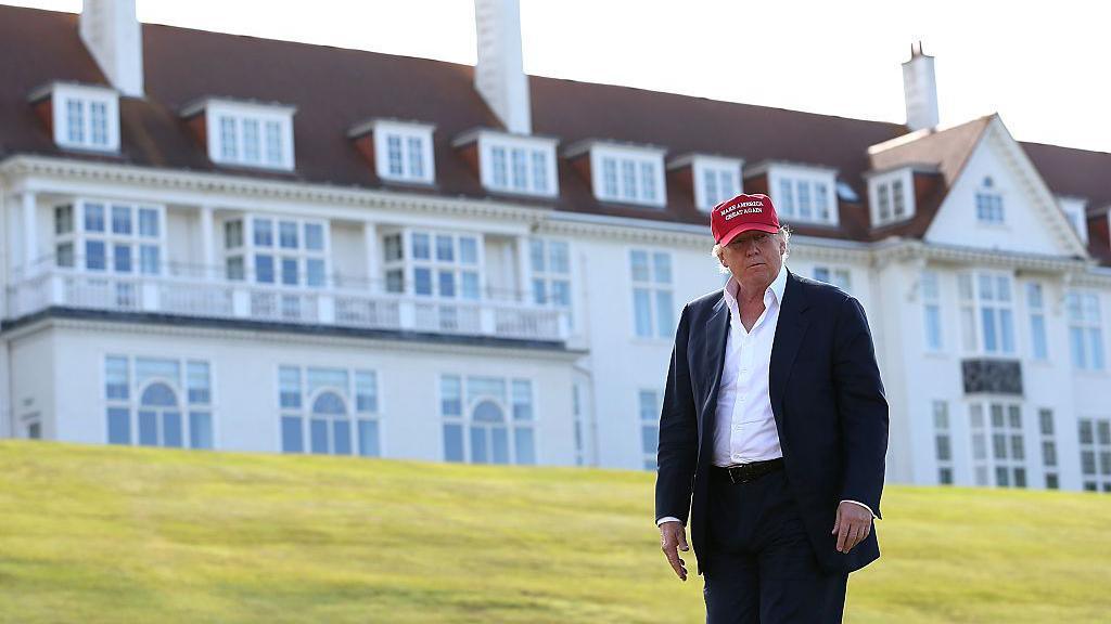 Trump at Turnberry. He is standing outside the hotel's famous white facade. He is wearing a dark suit, plain white shirt with no tie and a red 'Make America Great Again' baseball cap. 