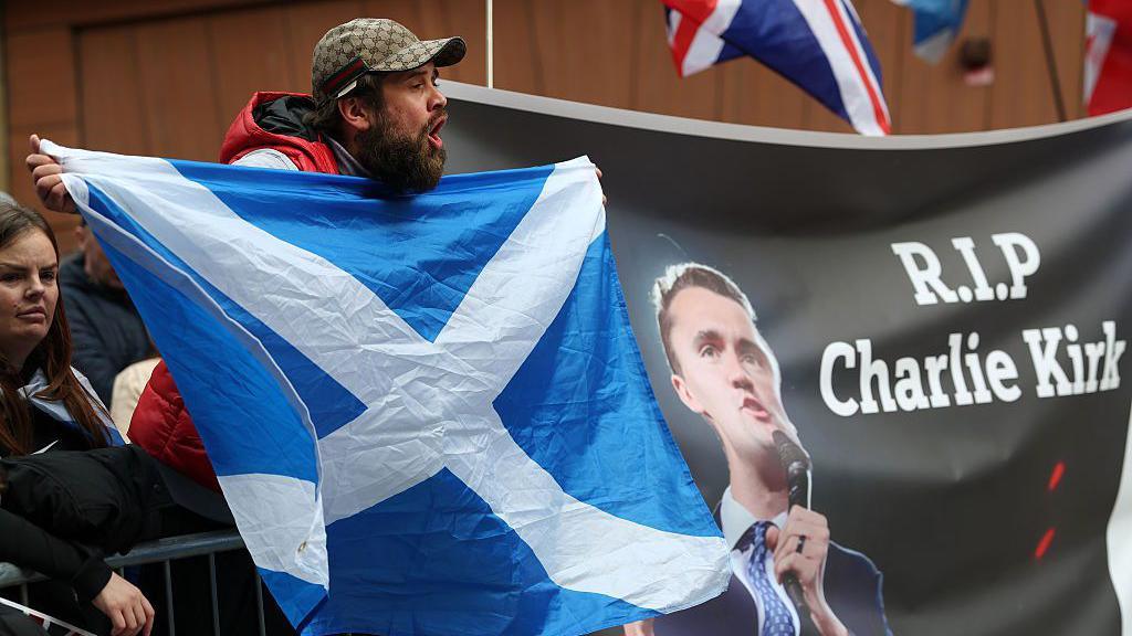 Demonstrators attend a rally on 20 September in Glasgow. A man with a beard, wearing a baseball hat, is holding a saltire next to a poster which says RIP Charlie Kirk.