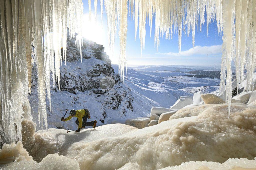 A climber holding an ice pick aloft is framed by icicles of a waterfall