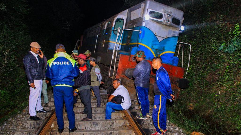 A group of people, including a man in a blue jacket with the word "police" printed on the back, stand on a railway track. Behind them is a derailed train.