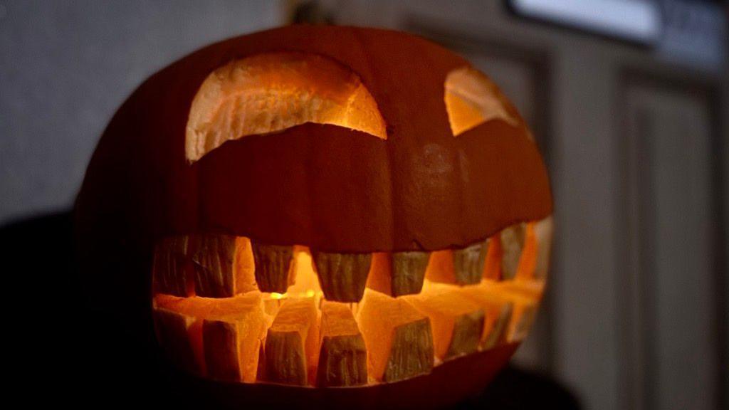 A carved orange pumpkin outside of a light brown door. The pumpkin has semi circle eyes carved and a mouth with lots of rectangular teeth. It is lit up with candles inside 