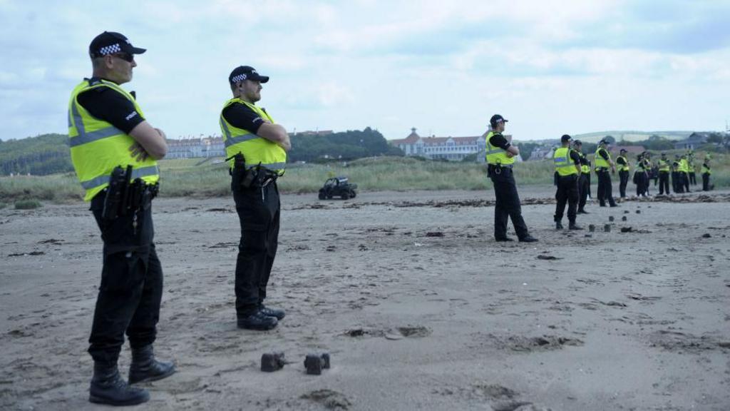 A line of Police Scotland officers on a beach  with the Turnberry golf course and hotel visible behind them.