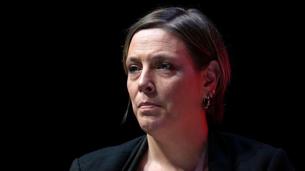 Jess Phillips has short brown hair and is sitting at an angle against a black background. Her head and shoulders are visible, and she is wearing a black suit jacket