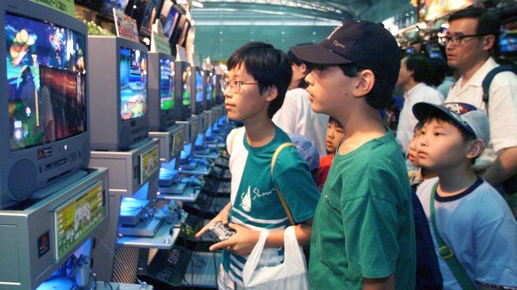 Several children gather around a series of screens, with one holding a controller and playing a video game. The PlayStation console is attached to the screen and outputting a game. It is an old CRT screen.