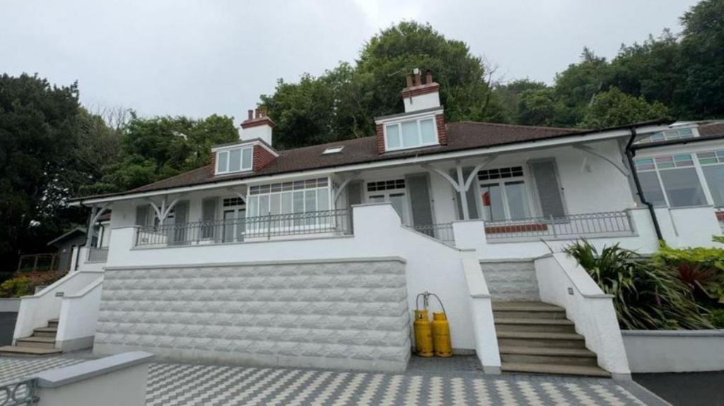 A white house with archways surrounding the front with steps either side leading to the verandah. There are two dormer windows on the first floor and trees behind it.