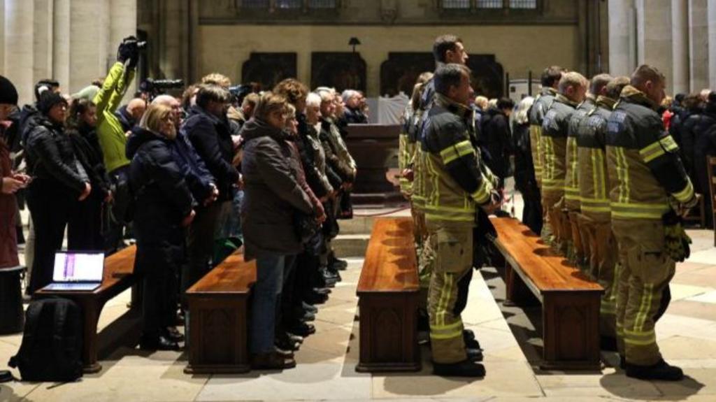 Members of the public and firefighters standing during Magdeburg Cathedral service. Members of the press can be seen in the back pews taking photos while attendants contemplate