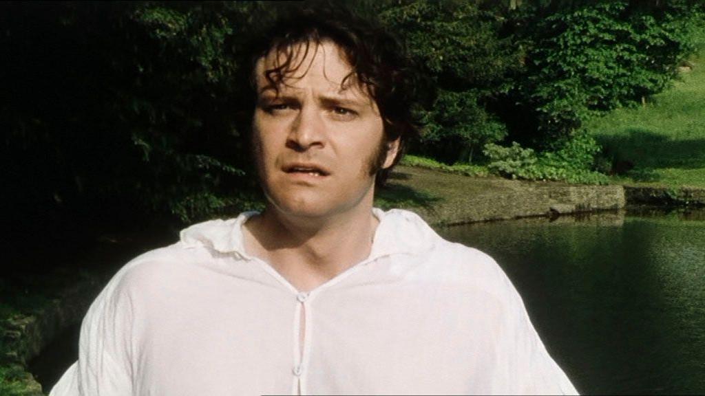 Colin Firth as Mr Darcy in the 1995 BBC drama Pride and Prejudice. He is wearing a white shirt while standing near a pond. Trees can be seen in the backdrop.