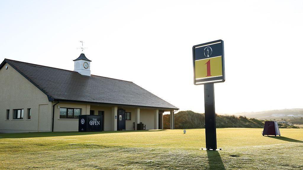 A large sign has the number '1' on it in red font. It's sits on a green grass tee box. Beside the tee box is a small cream coloured building. The building has a white clock on top of it.