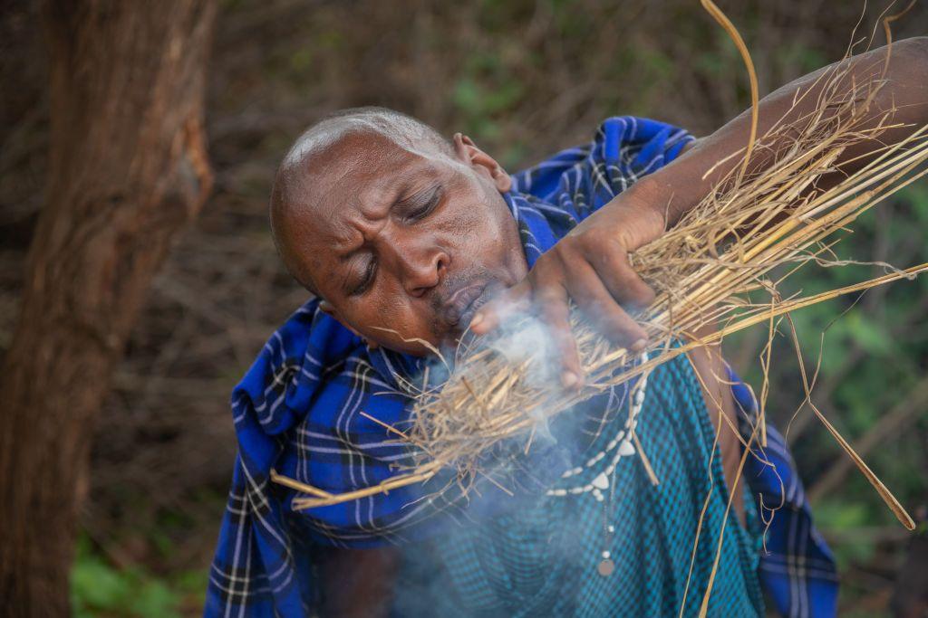 A man dressed in patterned blue clothing lights kindling and blows on it to accelerate the flame.