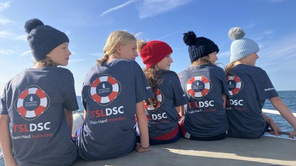 A line of girls on a boat, with their backs to the camera and wearing wooly hats, with tops on that say 'TSLDSC' and 'Train hard, swim easy'