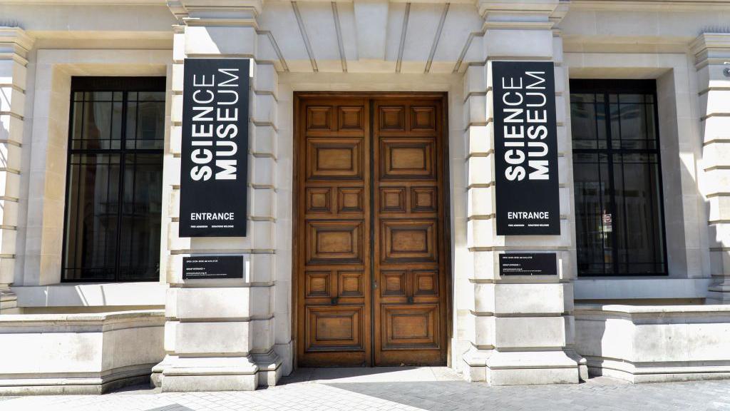 An external view of the Science Museum's entrance - two large wooden doors in a stone building, with black and white vertical signs on pillars either side