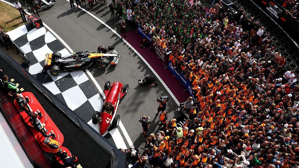 A shot from above of the podium at the British Grand Prix. A Formula One car is visible alongside a classic red F1 car. A large group of people are to one side looking towards the podium.
