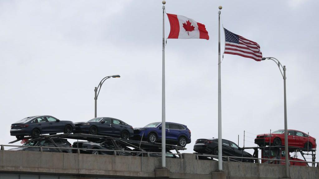 The Canada and US flags flap in the wind on flag polls as a truck stacked with cars for sale drives by
