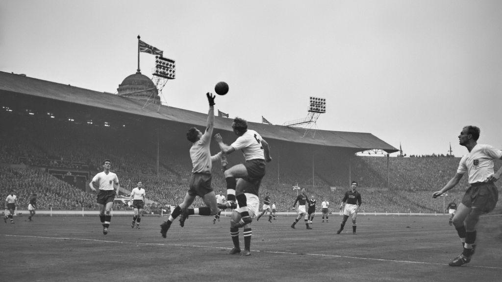 A black-and-white image showing England goalkeeper Colin McDonald leaping into the air with his right arm raised as he challenges for the ball during a friendly match at Wembley Stadium in october 1958. He is wearing a light, plain jersey, dark shorts and socks, as well as a pair of dark gloves. England's number 5 is also jumping for the ball. Thousands of people can be seen in the background in the stadium's stands. A union jack flag is hanging from one of the ground's towers.