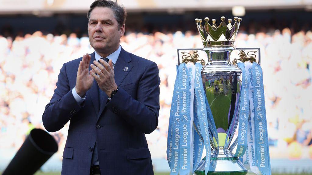 Richard Masters standing next to the Premier League trophy, which is in Manchester City colours