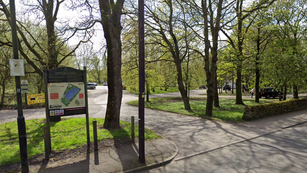 The entrance to Tarnfield Park in Yeadon. A cluster of trees stands either side of a road into the park, with cars dotted around in the background.