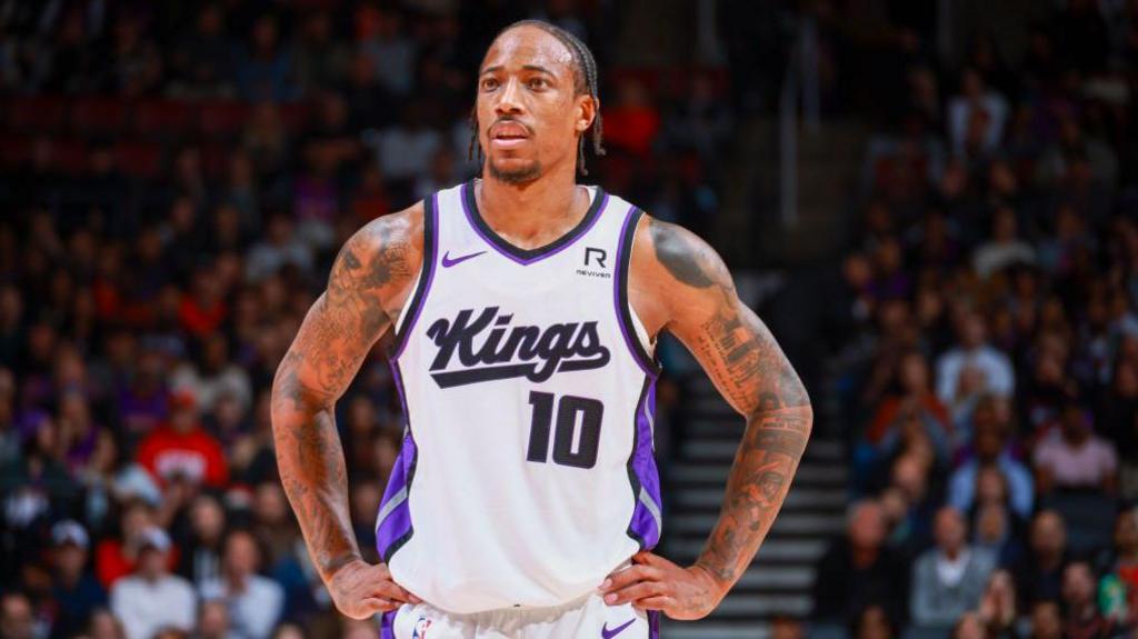 A basketball player in a white jersey with "Kings" and "10" printed on it stands in the middle of a court, resting his hands on his hips during a break in play. His forehead and heavily tattooed arms glisten with sweat.