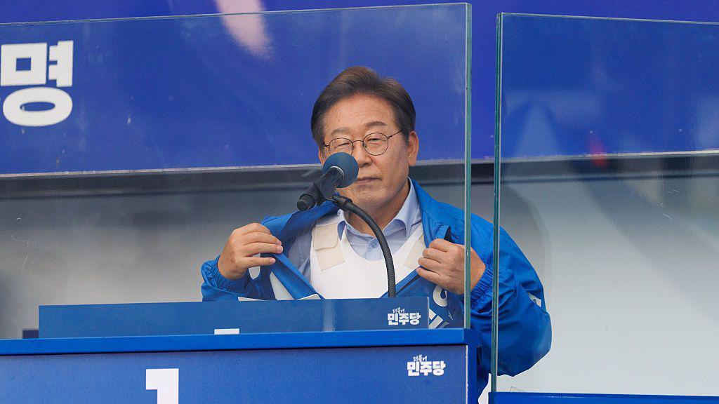 Standing at a podium behind bulletproof glass, Lee Jae–myung pulls open a blue tracksuit jacket to show a bulletproof vest underneath
