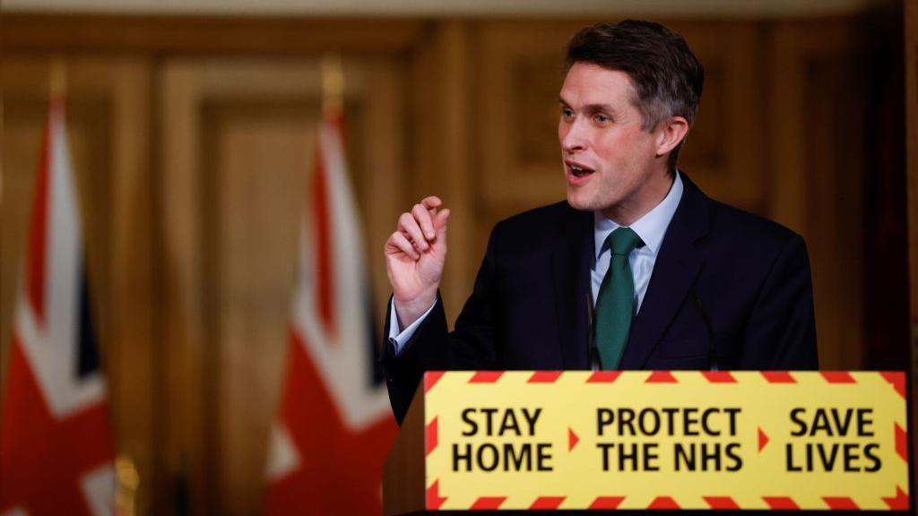 Gavin Williamson talks behind a podium that says STAY HOME, PROTECT THE NHS, SAVE LIVES. He is wearning a green tie, white shirt and black suit.
