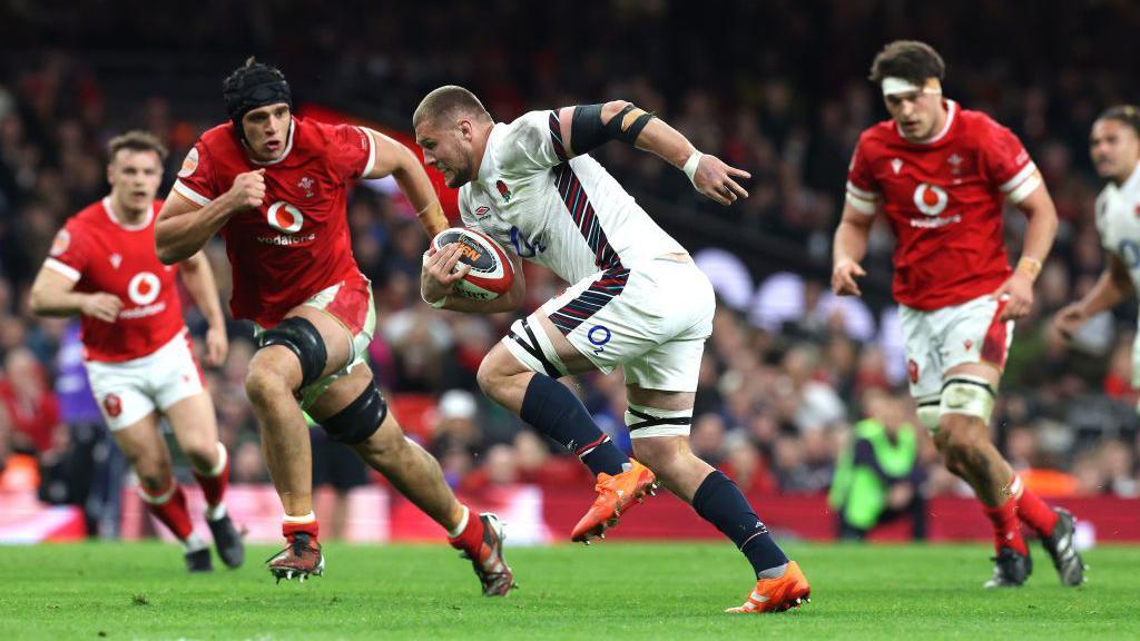 england player Tom Willis in a white kit holding the rugby ball surrounded by welsh players trying to get it off him 