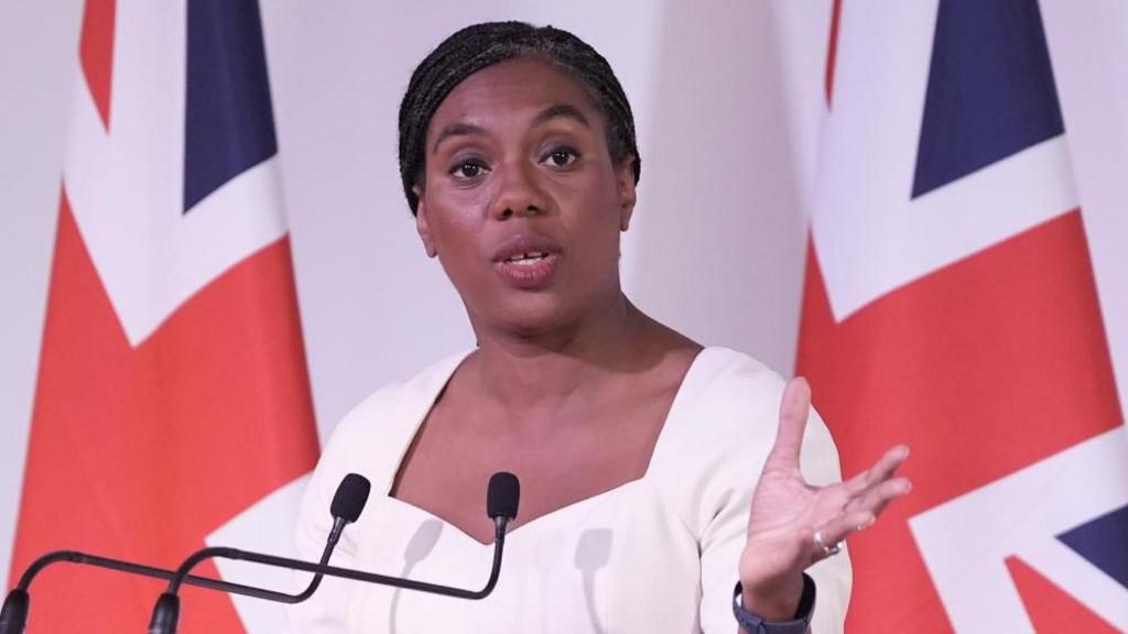 Kemi Badenoch gestures with her hand. She wears a white dress and stands in front of a union jack flag.