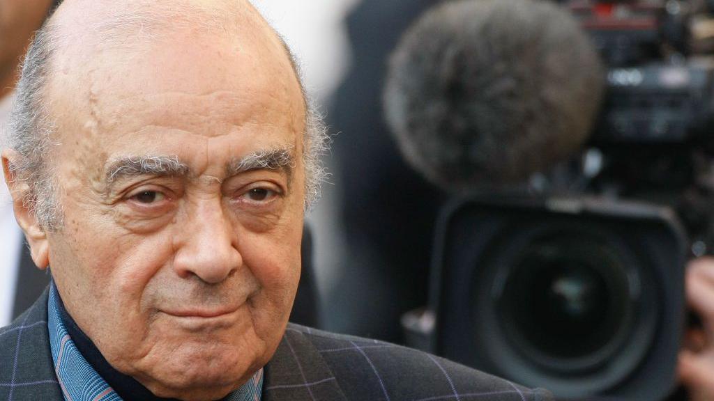 Mohamed Al Fayed with mild smirk. He is looking straight on. His eyebrows are fairly bushy and he is balding on the top of his head. He is wearing a black suit jacket with a thin checked pattern on. The photo was taken in 2008.