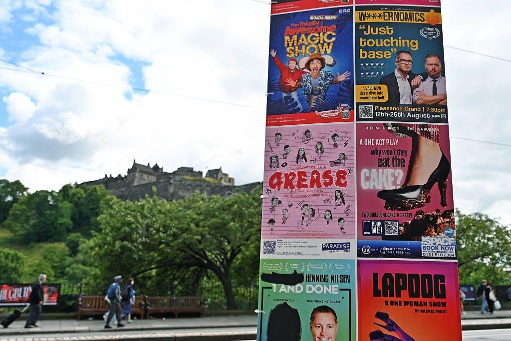 Brightly coloured posters advertising shows are pasted on a board with Edinburgh Castle in the background and the green trees of Princes Street Gardens below.