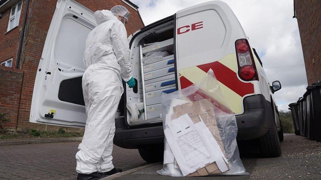 The back of a white police van with one of the back doors open. There is a person dressed in a white protective full body suit next to the open van door with their back turned to the viewer. The van appears to have storage in it and there is a clear bag of evidence outside the van on the floor.