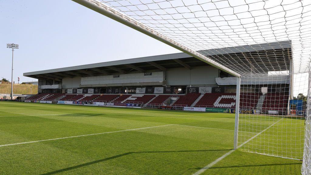 A general view of the East Stand at Sixfields Stadium. Photo is taken from inside the net on the football pitch looking towards the stands. The seats under the stand are mostly red. Some of the seats have been painted white to spell out NTFC. The sky is blue with no clouds and to the left of the photo is floodlight, which has not been turned on. The pitch is well kept and square patches of grass have been carved out, they can be noticed as each square is a slightly different shade of green.