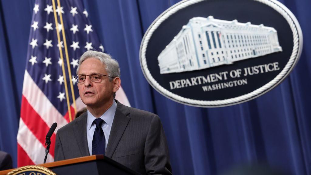 US Attorney General Merrick Garland, a man with white hair and glasses, stands before a lectern. An American flag and sign that says "Department of Justice, Washington" are behind him.