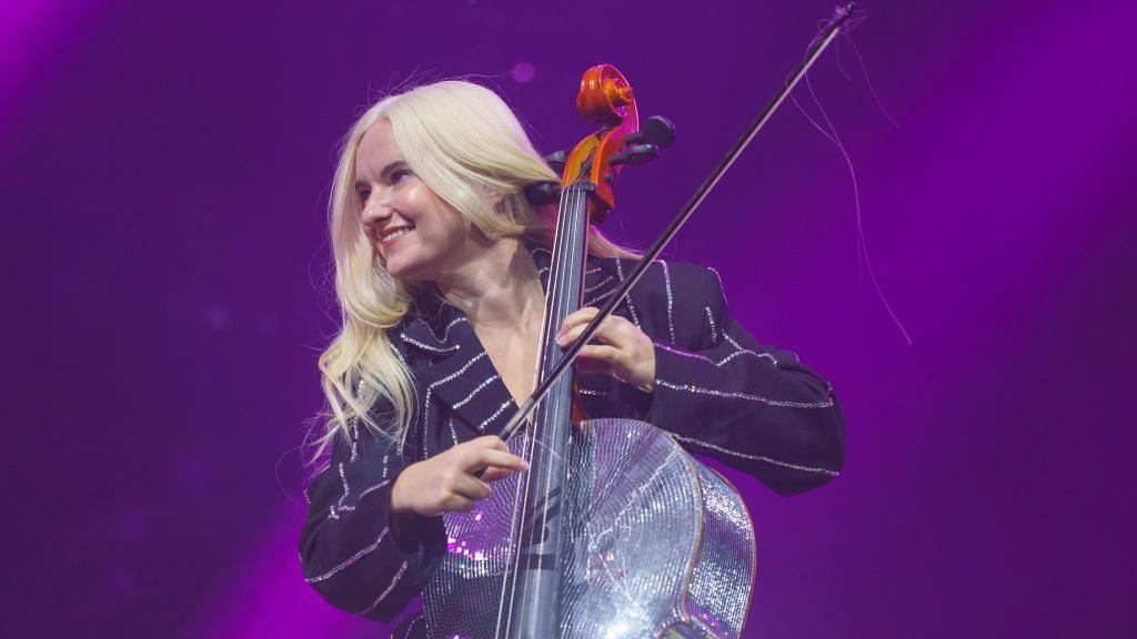 A woman with long blonde hair is on a stage. She is holding a silver glittery cello and a bow. The light behind her is purple
