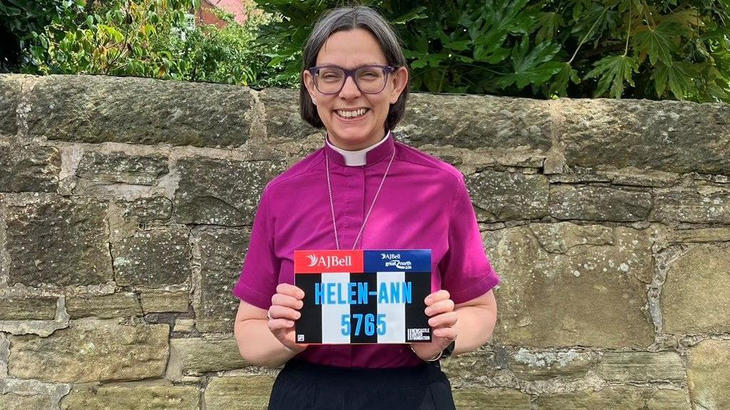 The Bishop of Newcastle is standing in front of a wall wearing the purple shirt and dog collar of a bishop and a dark skirt. She has greyish brown hair and glasses and is in her 50s. She is holding up a number for the Great North Run 