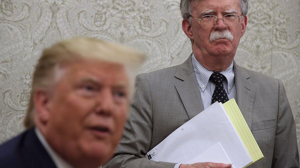 John Bolton, holding documents and a yellow legal pad, stands behind Donald Trump, who is seated, in the Oval Office. 