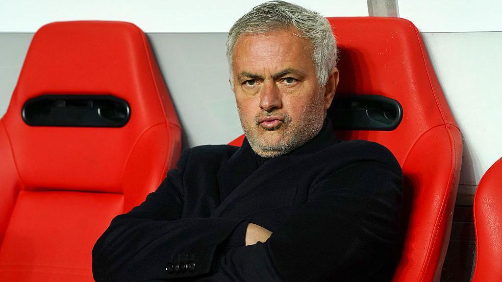Jose Mourinho purses his lips and cross his arms as he sits on a red chair