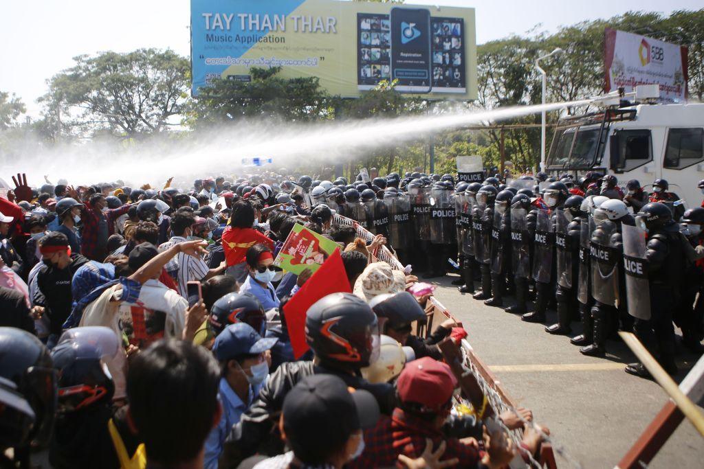 A line of police with riot shields and helmets face a crowd of protesters with a gap of a few metres between the two sides. Behind the police is a white vehicle spraying water from a water cannon on its roof, as protesters demonstrate against the 1 Feb military coup in the Myanmar capital Naypyidaw on 9 Feb, 2021