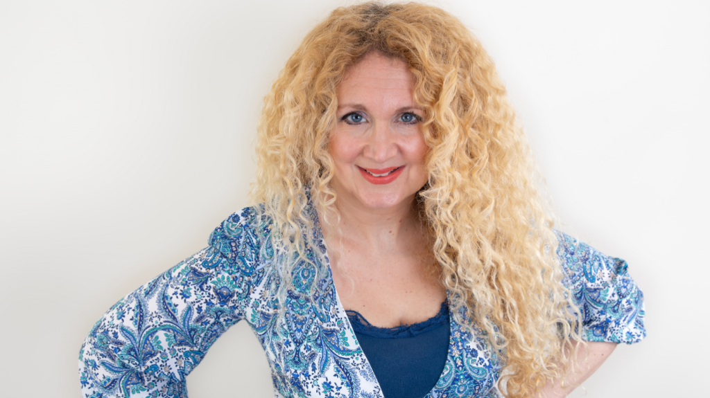 Woman with curly blonde hair is smiling. She's wearing a paisley blue and whict blouse and a dark blue camisole. 