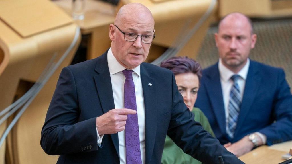 First Minister John Swinney stands during backbench FMQs with Angela Constance and Neil Gray sitting in the background