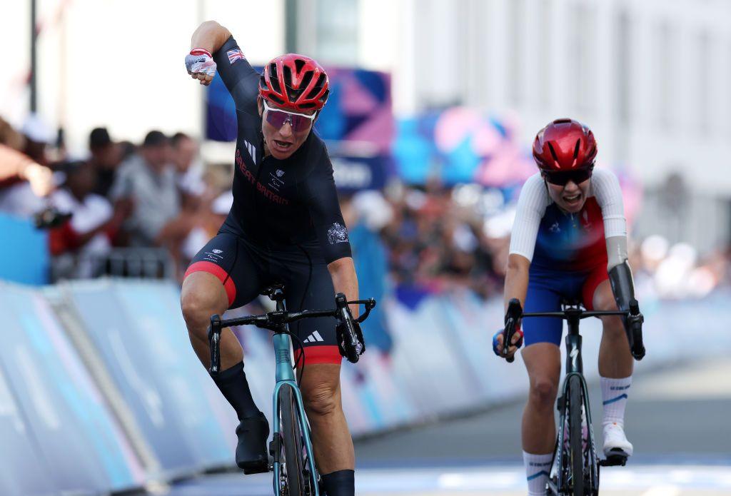 sarah storey lifts her arm in the air while riding her bike to the finish line as her competitor is just moments behind her on the track