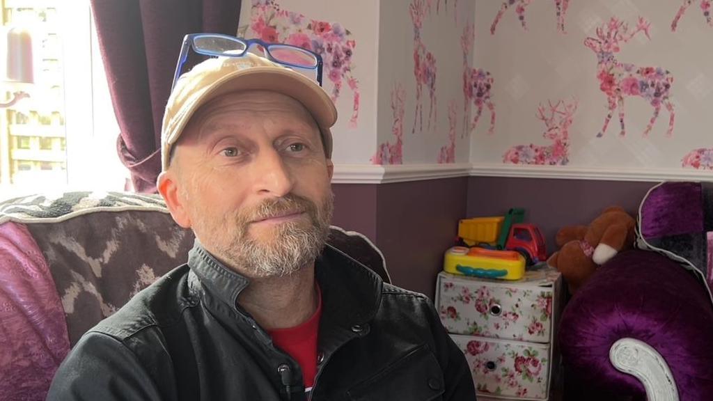 Al Coates has a beard and is wearing a baseball cap with glasses perched on the top of his hat. He's sitting on a sofa in a front room with children's toys behind him and wallpaper with pink and purple reindeer on a white background.