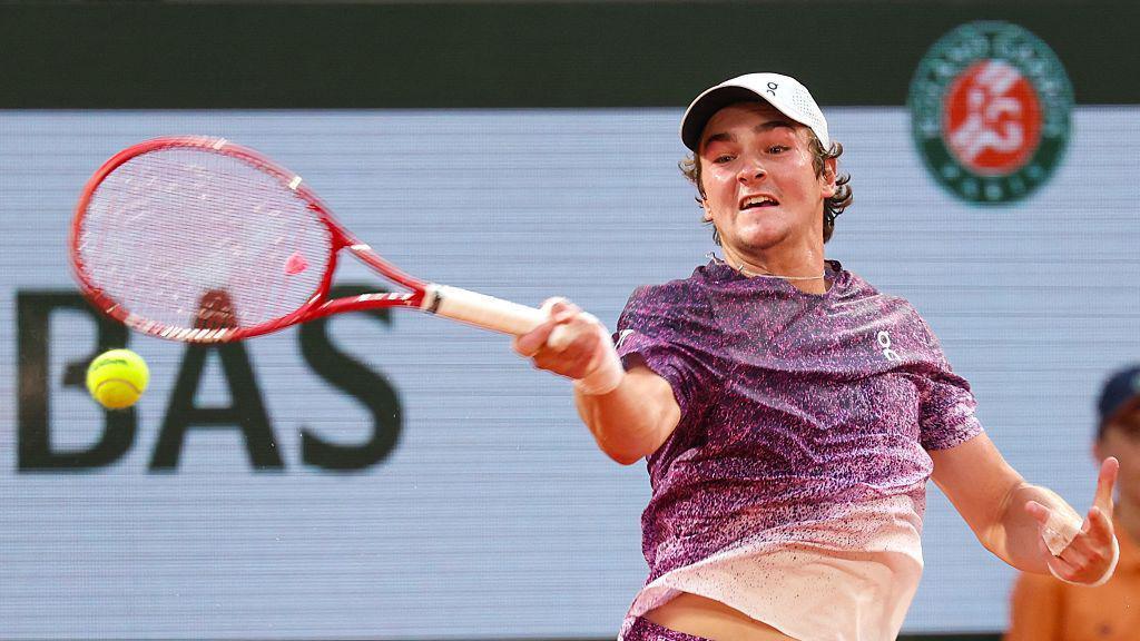 Joao Fonseca, wearing a purple t-shirt which fades into white and a white cap, plays a forehand with his red racquet in his right hand and the yellow ball just below it. 