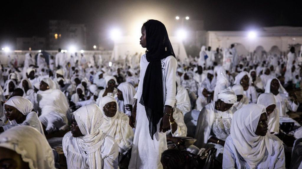 An image of a young woman standing in front of bright white light creating a halo-like effect, surrounded by other women who are sitting down and dressed in fully white garments in Dakar, Senegal - Sunday 15 September 2024.