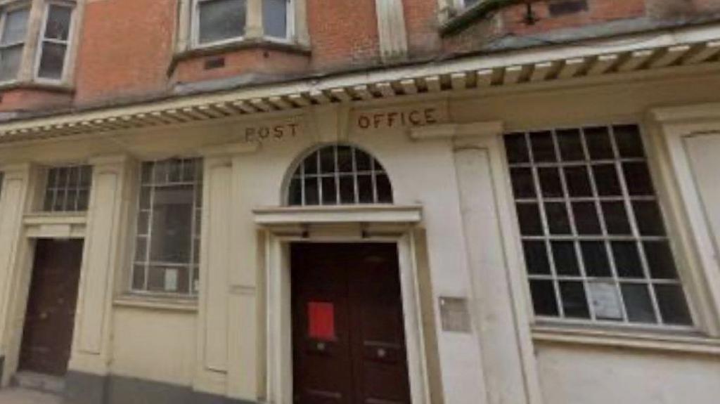 The front of an old post office. The building is white with a brown door. The building says POST OFFICE on it in red letters. There are windows on either side of the main door. To the left of the window on the left of the door is another smaller door. Above the white part of the building is red brick and windows. 