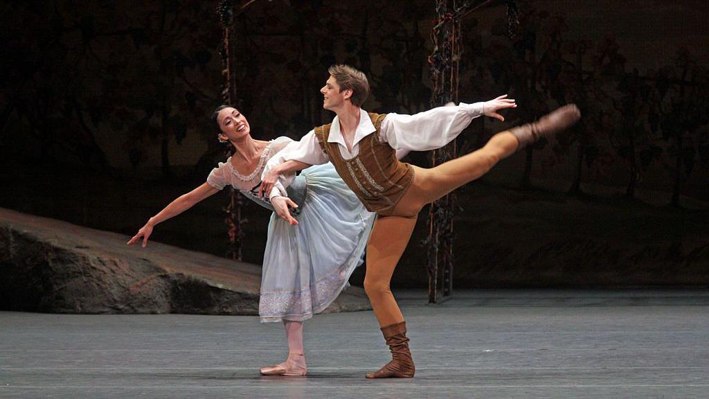 Two ballet dancers perform Giselle. The male dancer wears a brown vest and boots, executing a dramatic move while supporting his partner, who wears a light blue peasant dress with white sleeves. She leans back in an expressive pose with her arms gracefully extended. They are performaning on a dimly lit stage with a rustic set design.