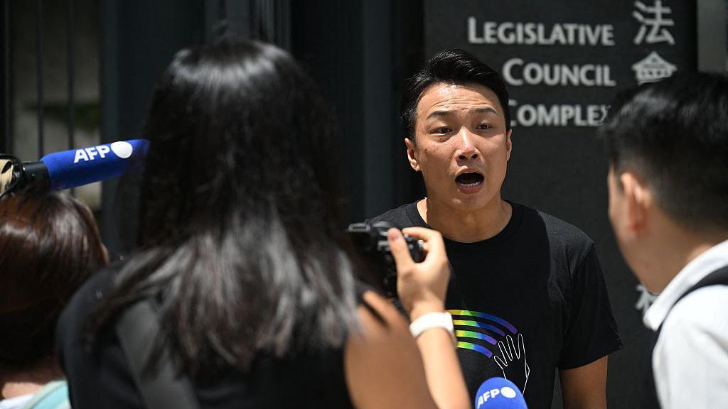 Jimmy Sham wearing a black shirt with a rainbow logo speaks to reporters outside of the Legislative Council Complex in Hong Kong
