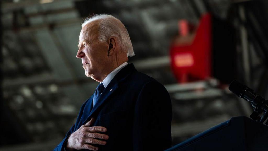 Former US President Joe Biden's side profile. His hand is laid on his chest and he has a dark coat on.