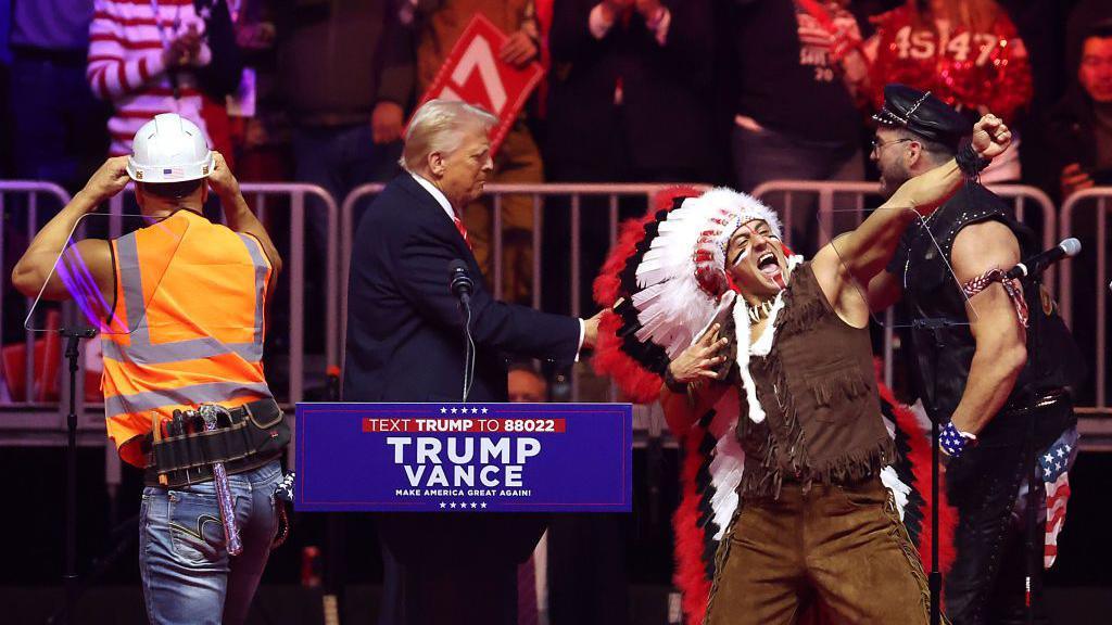 Donald Trump appears on stage as members of the Village People, wearing a Native American headdress and construction gear, preform around him. 