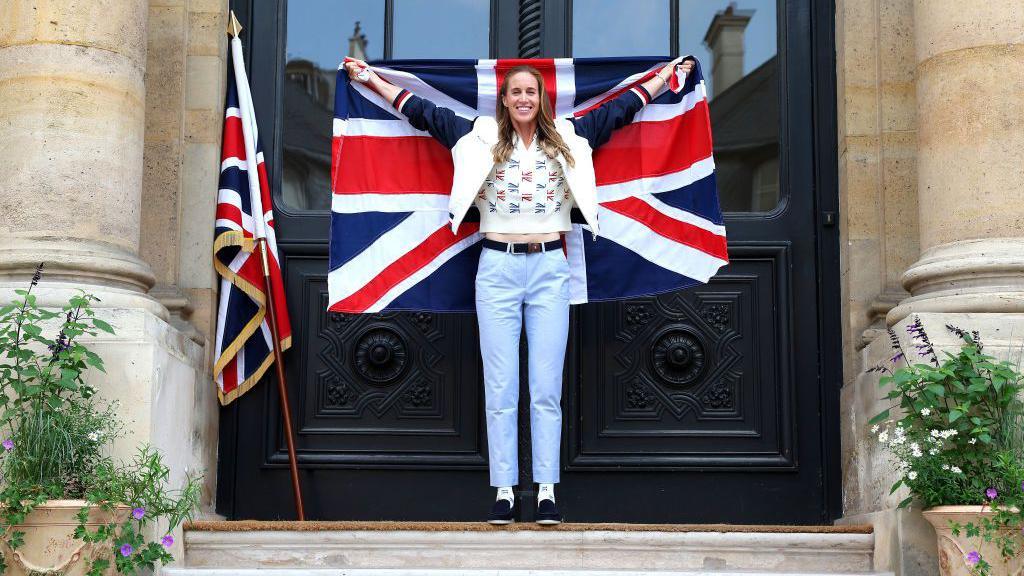 A woman with long brown hair wearing blue jeans and a white top, standing outside a pair of smart-looking black doors holding up a large Union Jack and smiling.