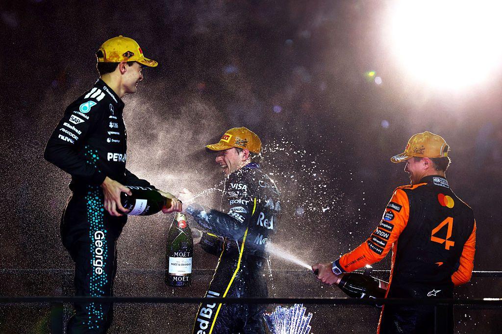 Three Formula 1 drivers celebrate on the podium, spraying champagne from large bottles. They wear racing suits and gold caps, with bright lights illuminating the scene and champagne mist filling the air against a dark background.
