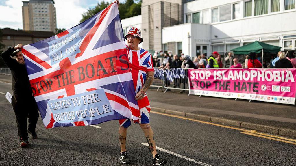 Anti-migrant protesters outside the Cladhan Hotel on 13 September. Two men carry a union flag with the phrase Stop the boats written on it. The man at the back is dressed in black and the man at the front is wearing shorts, a tshirt and a hat which all feature the flag. In the background are counter-protesters and a banner reading: Stop Islamophobia. Stop the hate.