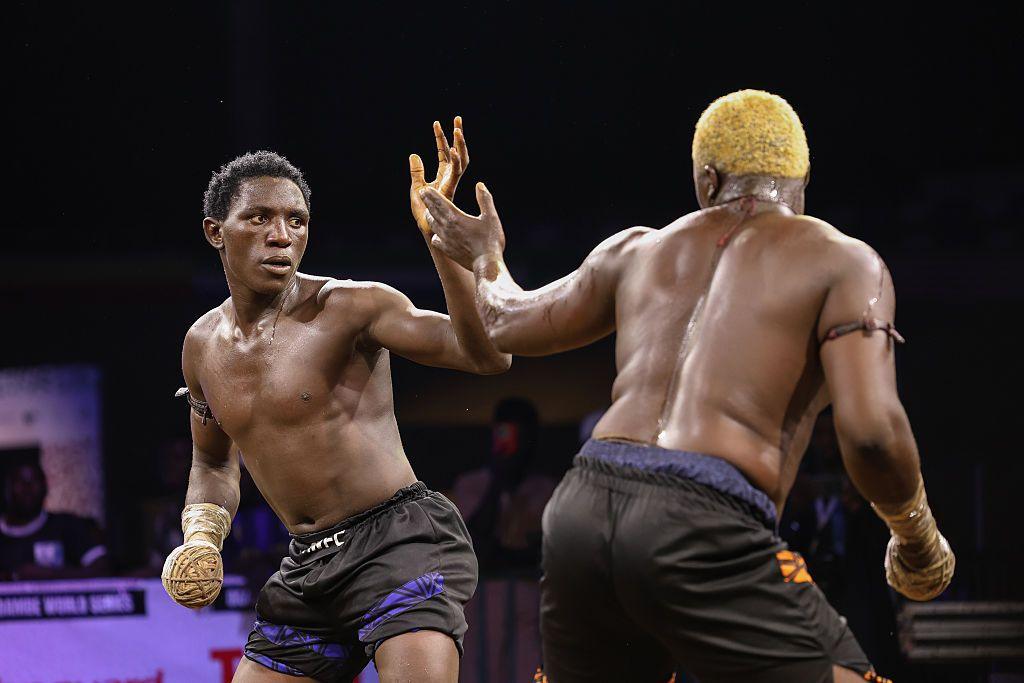 Two men square up to each other in an area. Both are dressed in shorts, both have right hands balled up and taped into a hard fist.
